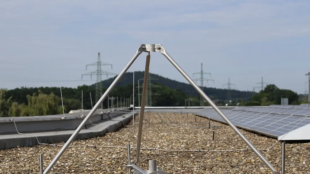 Metal tripod structure mounted on concrete blocks on a gravel rooftop, with solar panels and power lines visible in the background.