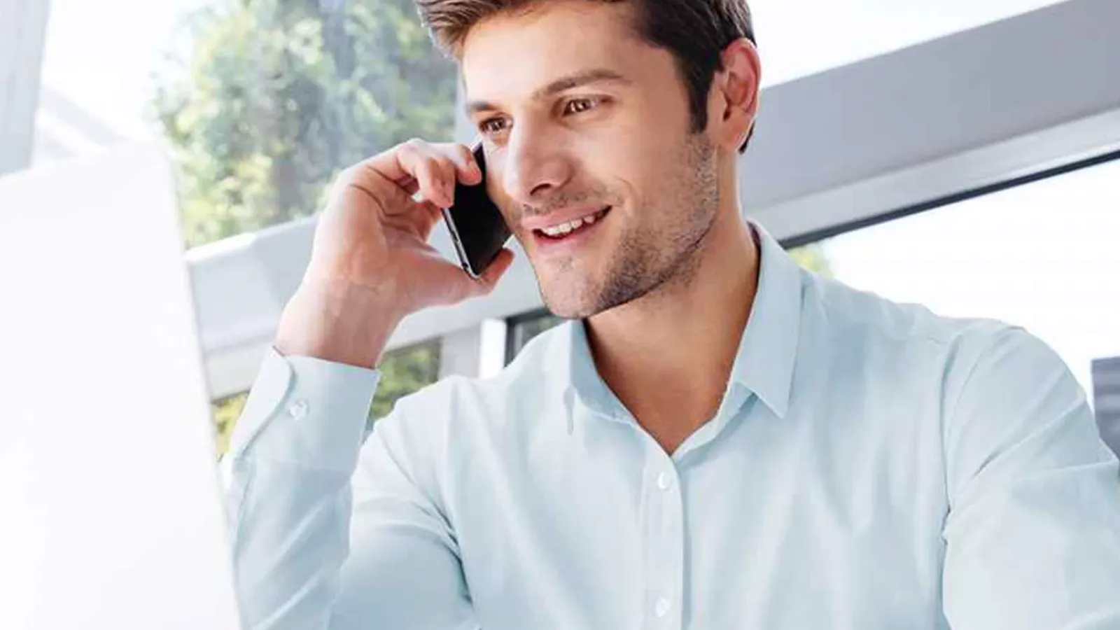 Man in a light blue shirt talking on a smartphone while working on a laptop in a bright office setting.