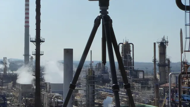 A tripod-mounted measuring instrument is aimed at a large-scale industrial plant with chimneys, pipelines, and silos.