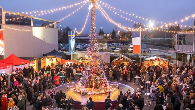 Une scène festive de marché de Noël met en scène un grand sapin illuminé, orné de lumières et de décorations. Une foule de personnes vêtues de tenues d'hiver se rassemble autour des stands festifs, profitant de l'ambiance animée et joyeuse. Des guirlandes