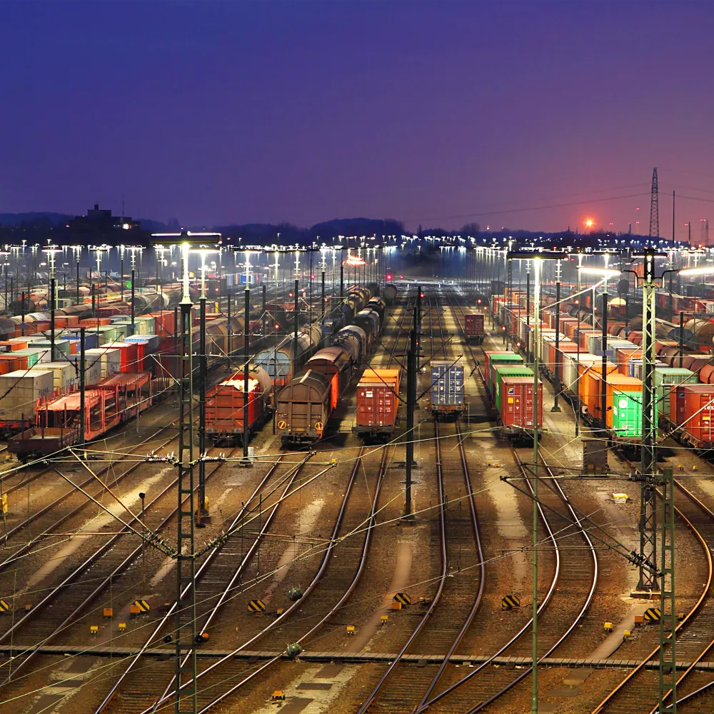 Numerous cargo trains and containers are lined up in a brightly lit train yard at dusk, with rail tracks converging into the distance and the sun setting on the horizon.