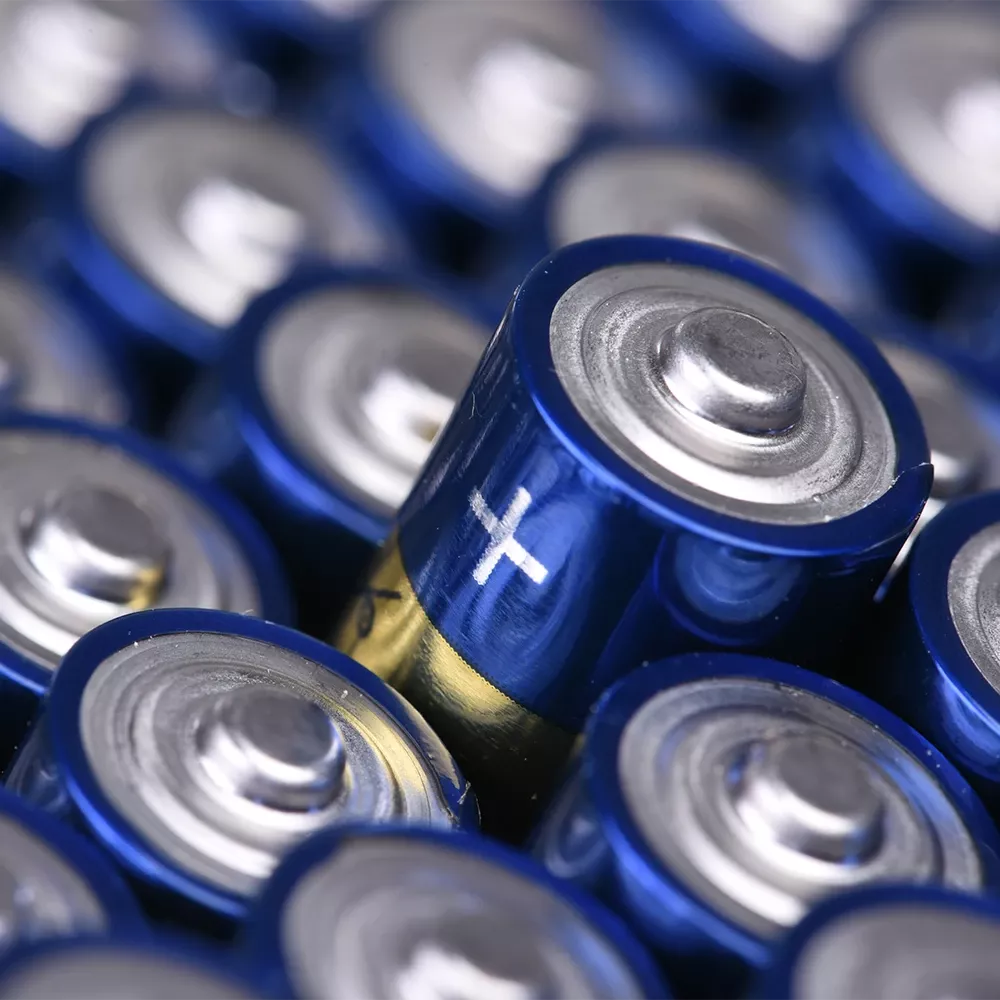 Close-up of multiple blue cylindrical batteries, with one battery marked with a white plus symbol indicating the positive terminal.