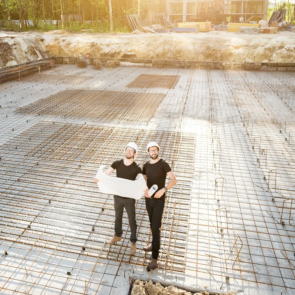 Two construction workers wearing hard hats stand on a reinforced concrete foundation slab, holding blueprints at a building site.