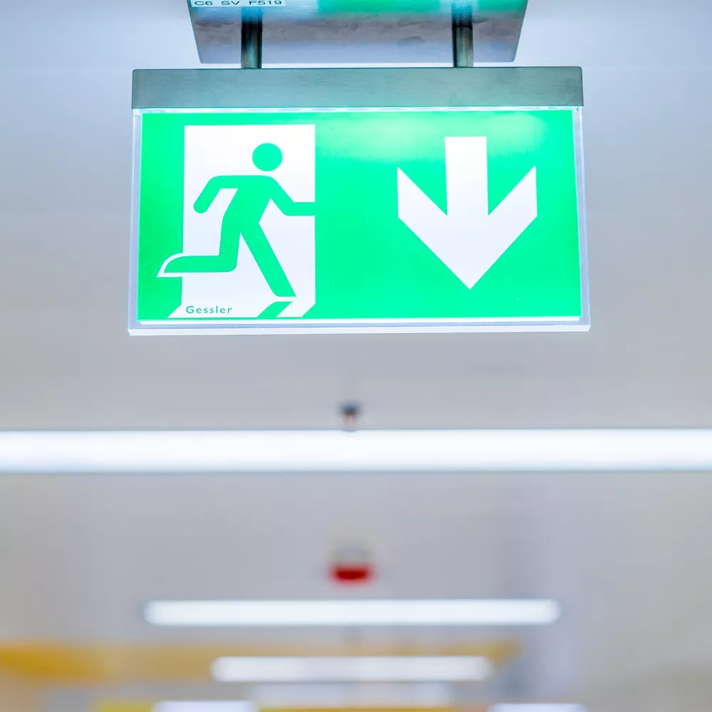 Illuminated green emergency exit sign with a running figure and a downward arrow, mounted on a ceiling in an indoor corridor.