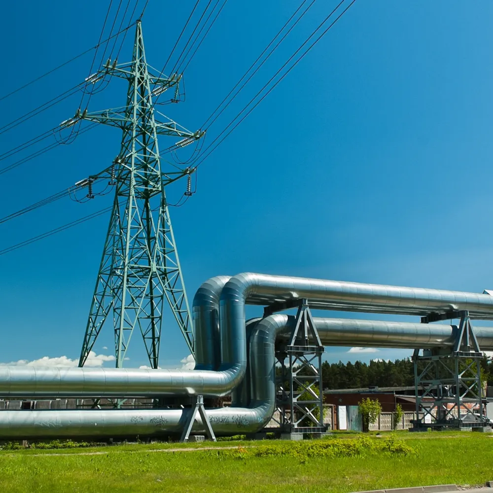 Industrial pipeline installation beneath a high-voltage transmission tower with overhead power lines under a clear blue sky.