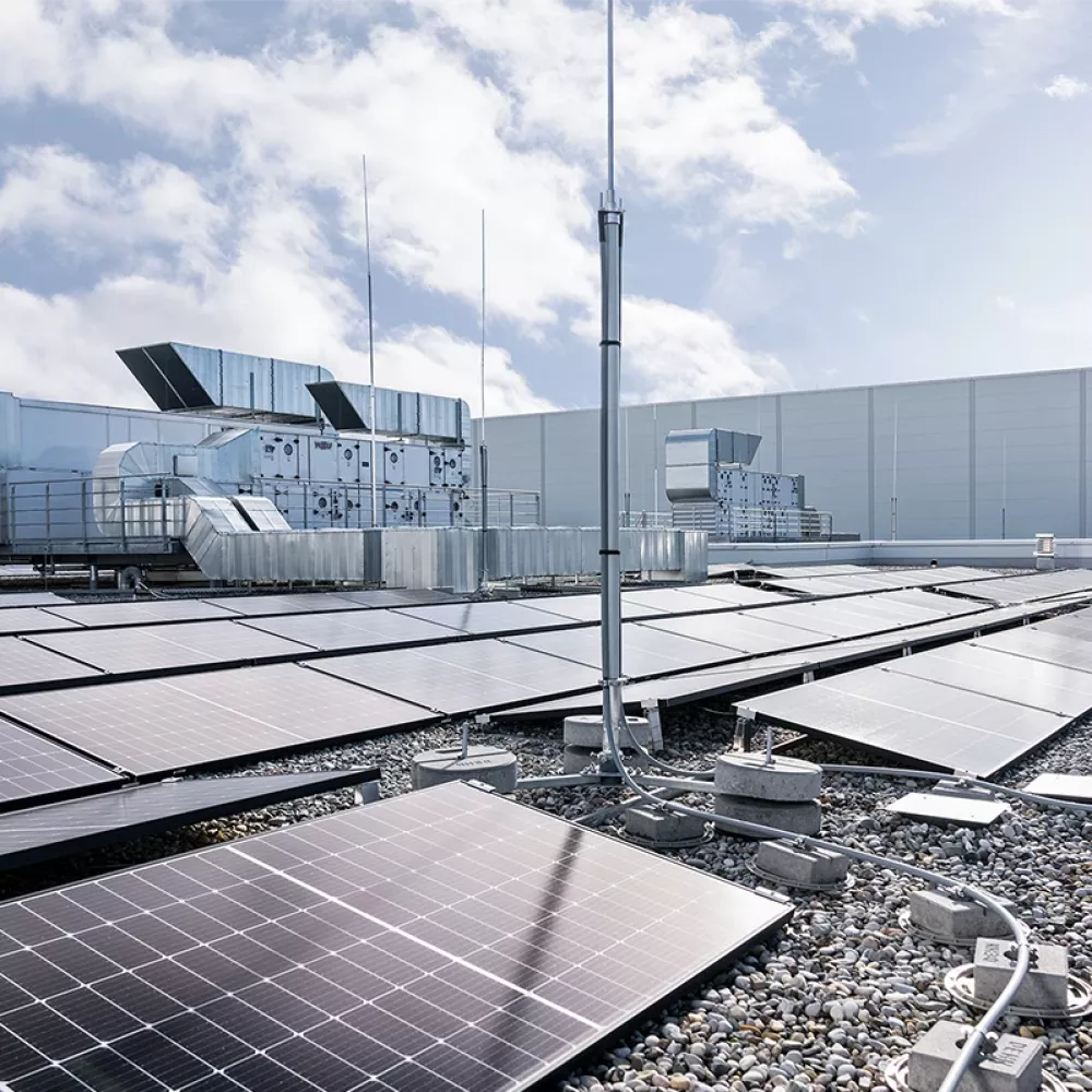 Rooftop photovoltaic system with solar panels, lightning protection mast, and cable routing on a commercial building roof under a cloudy sky.
