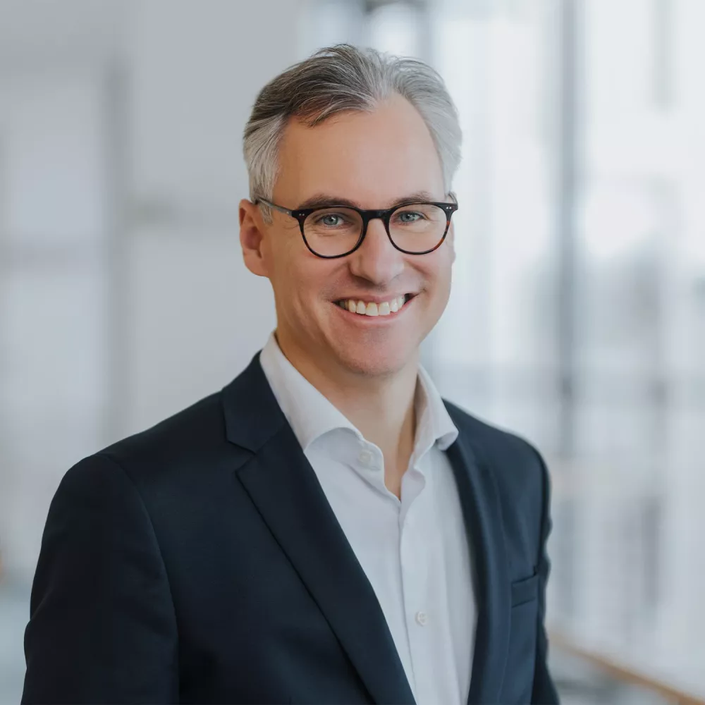 Portrait of a smiling man with gray hair and glasses, wearing a dark suit jacket and white shirt, in a softly blurred indoor setting.