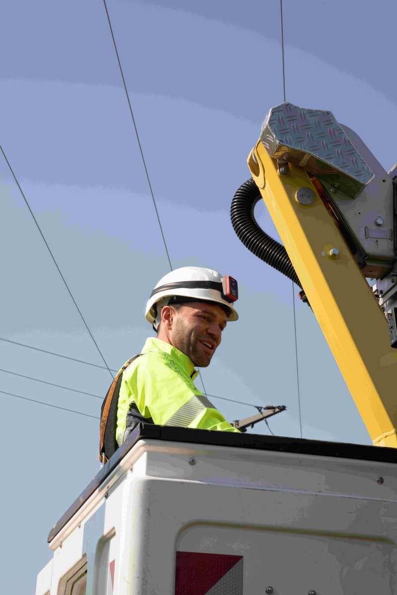 A captivating image displays a man in a white hard hat and yellow vest, expertly perched on a massive utility truck with a crane. His sunny smile and the trucks electrical machinery create a visually striking scene against a clear blue sky.