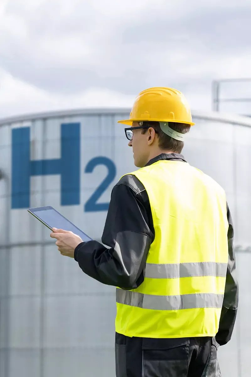 A captivating image displays a man in a yellow hard hat and vest, standing by an H2 cylinder. The images unique blend of a worker holding a tablet amidst a serene blue sky with clouds creates a peaceful construction scene.