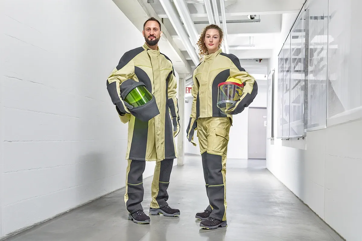 Two electricians in protective workwear stand in an industrial corridor, holding helmets with visors as part of modern personal protective equipment.