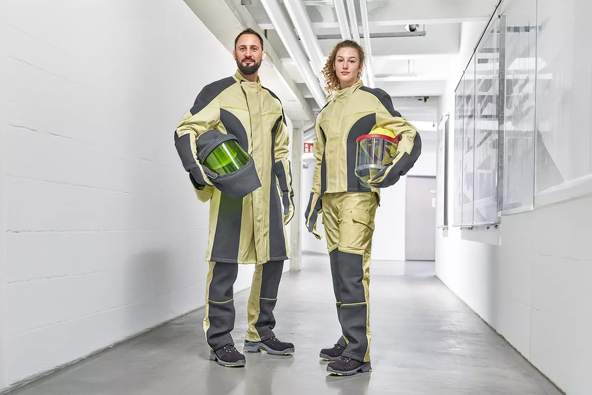 Two electricians in protective workwear stand in an industrial corridor, holding helmets with visors as part of modern personal protective equipment.