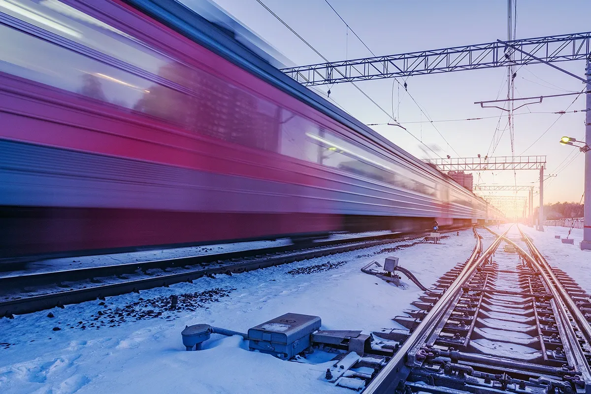 High-speed train passing a snow-covered railway junction at dusk, with blurred motion, overhead power lines, and visible switch components along the tracks.
