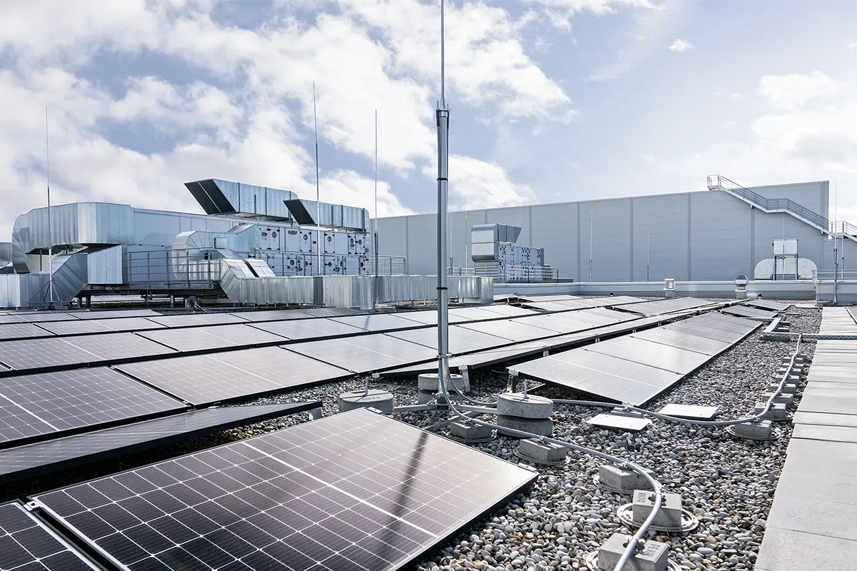 Rooftop photovoltaic system with solar panels, lightning protection mast, and cable routing on a commercial building roof under a cloudy sky.