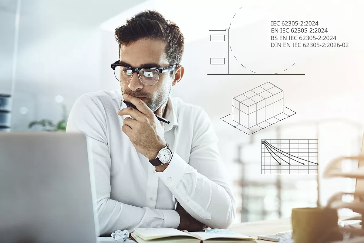 Man in glasses working at a laptop, holding a pen thoughtfully, with technical diagrams and IEC 62305-2:2024 graphics overlaid in the background.