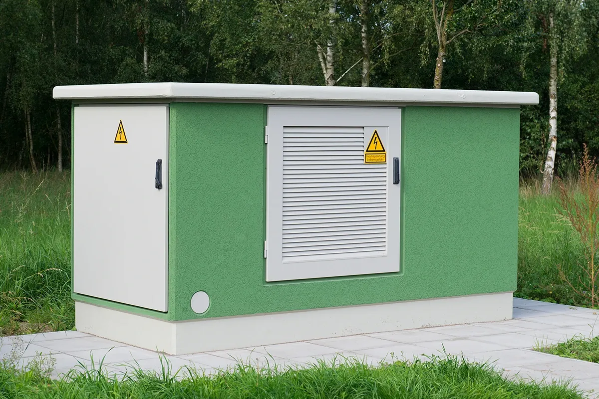 Green transformer station building with ventilation door and high-voltage warning signs, surrounded by grass and trees.