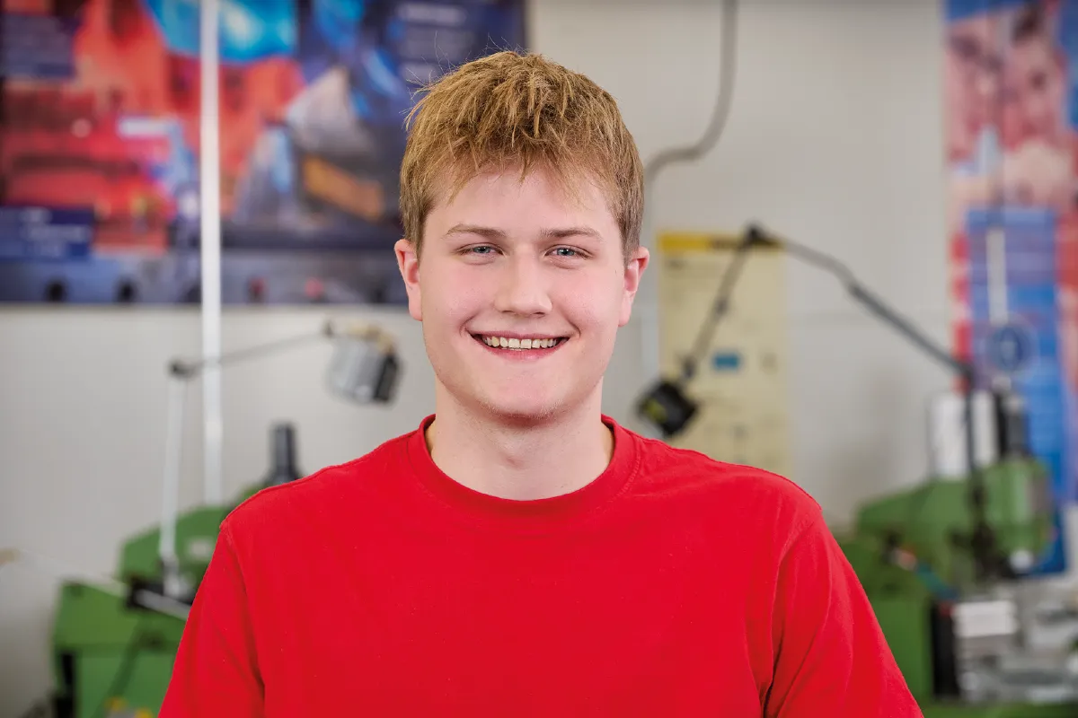 Person with short light hair wearing a red shirt in an industrial workshop with machinery in the background.
