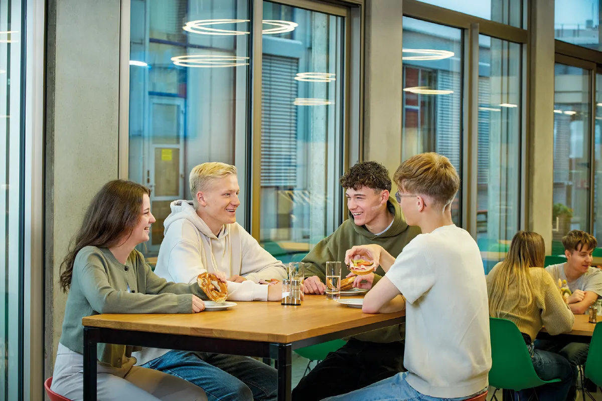 Group of young people sitting around a wooden table in a modern room with large windows and circular ceiling lights.